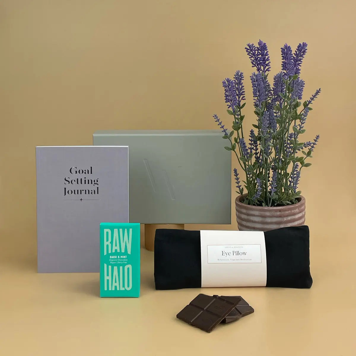 Selfcare Gift - Goal setting journal, raw halo product, eye pillow, chocolate, and lavender plant on a beige background